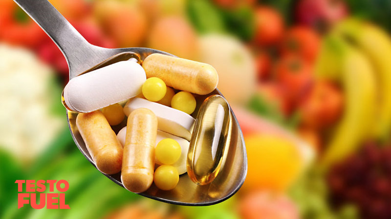 A range of different pills and tablet supplements on a spoon. Fresh vegetables and fruit in the background